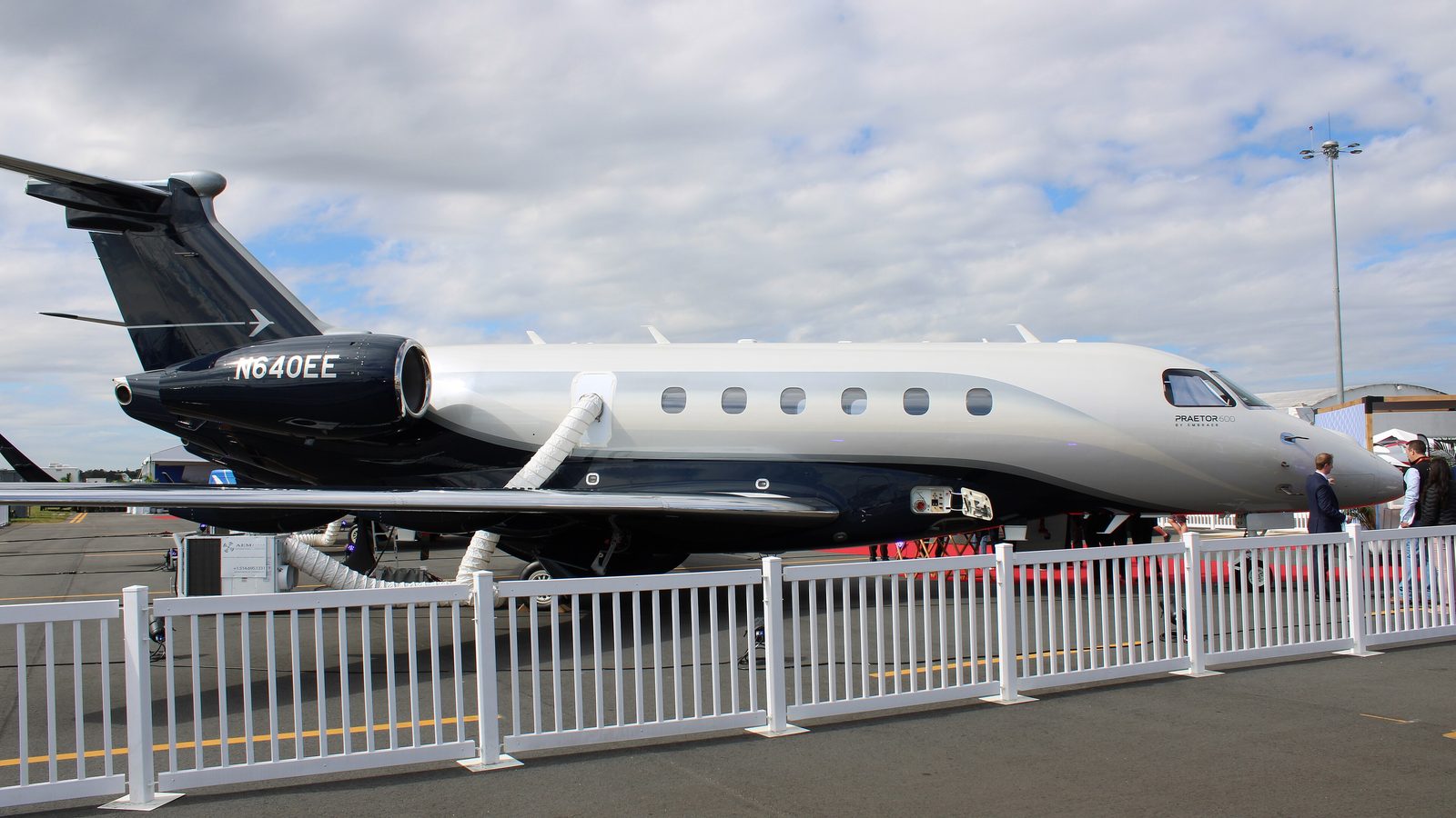 White and dark blue Embraer Praetor 600 on a static display ramp behind a white fence, with people standing at the nose