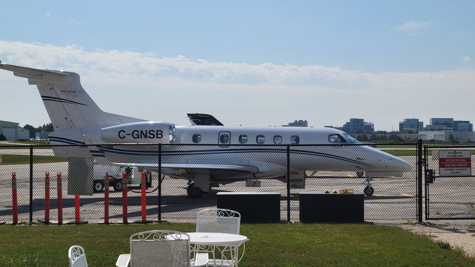 White Embraer Phenom 300 with blue stripes parked behind a chain-link fence, white garden chairs in the foreground