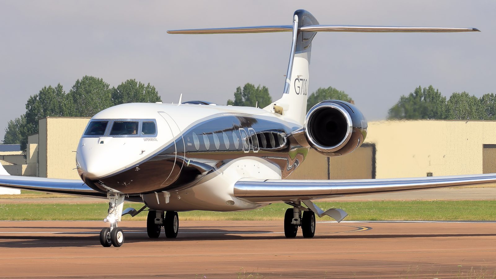 A white Gulfstream G700 with a dark grey belly on a taxiway, G700 on the tail, two rear engines, hangars and trees behind