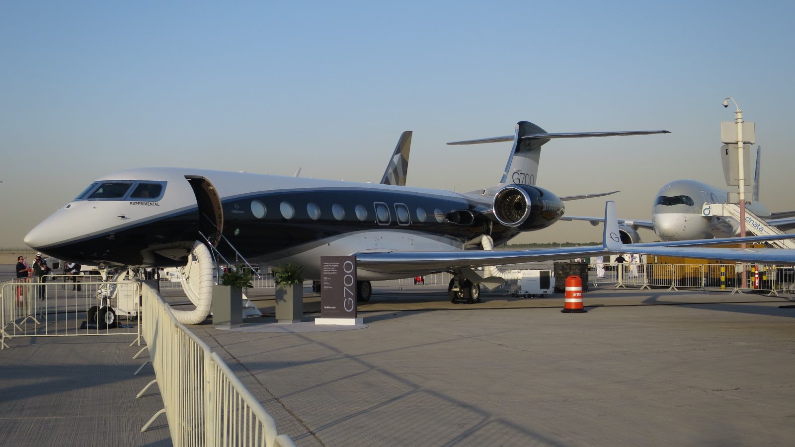A white and dark blue Gulfstream G700 on a static display behind a white fence, door open, G700 on the tail