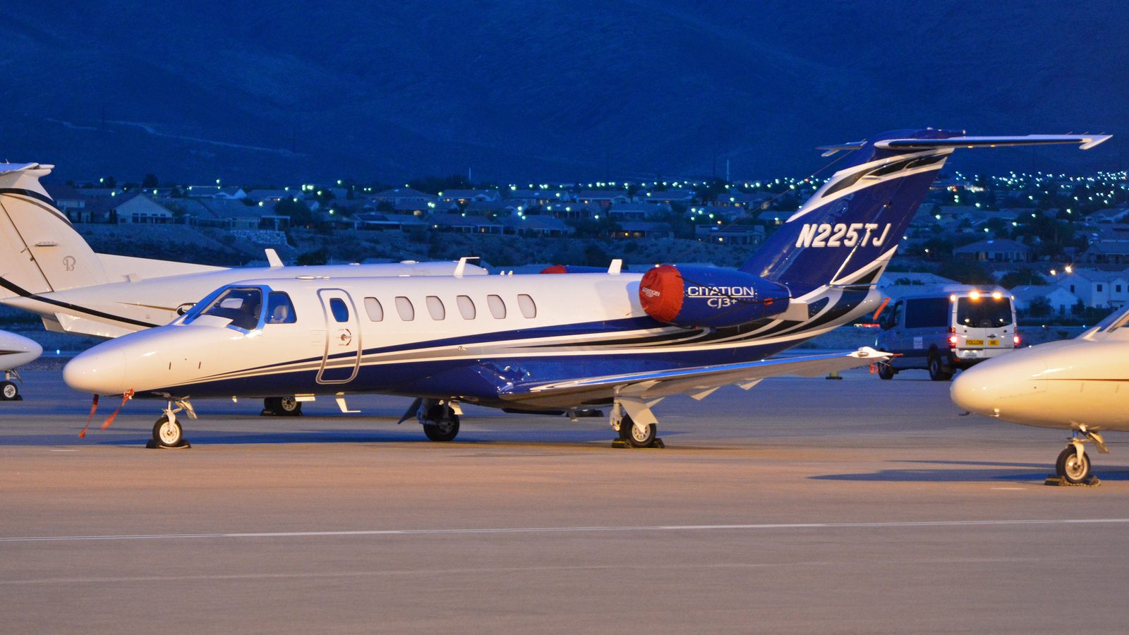 White and blue Cessna Citation CJ3+ on a ramp at dusk, with houses and hills behind