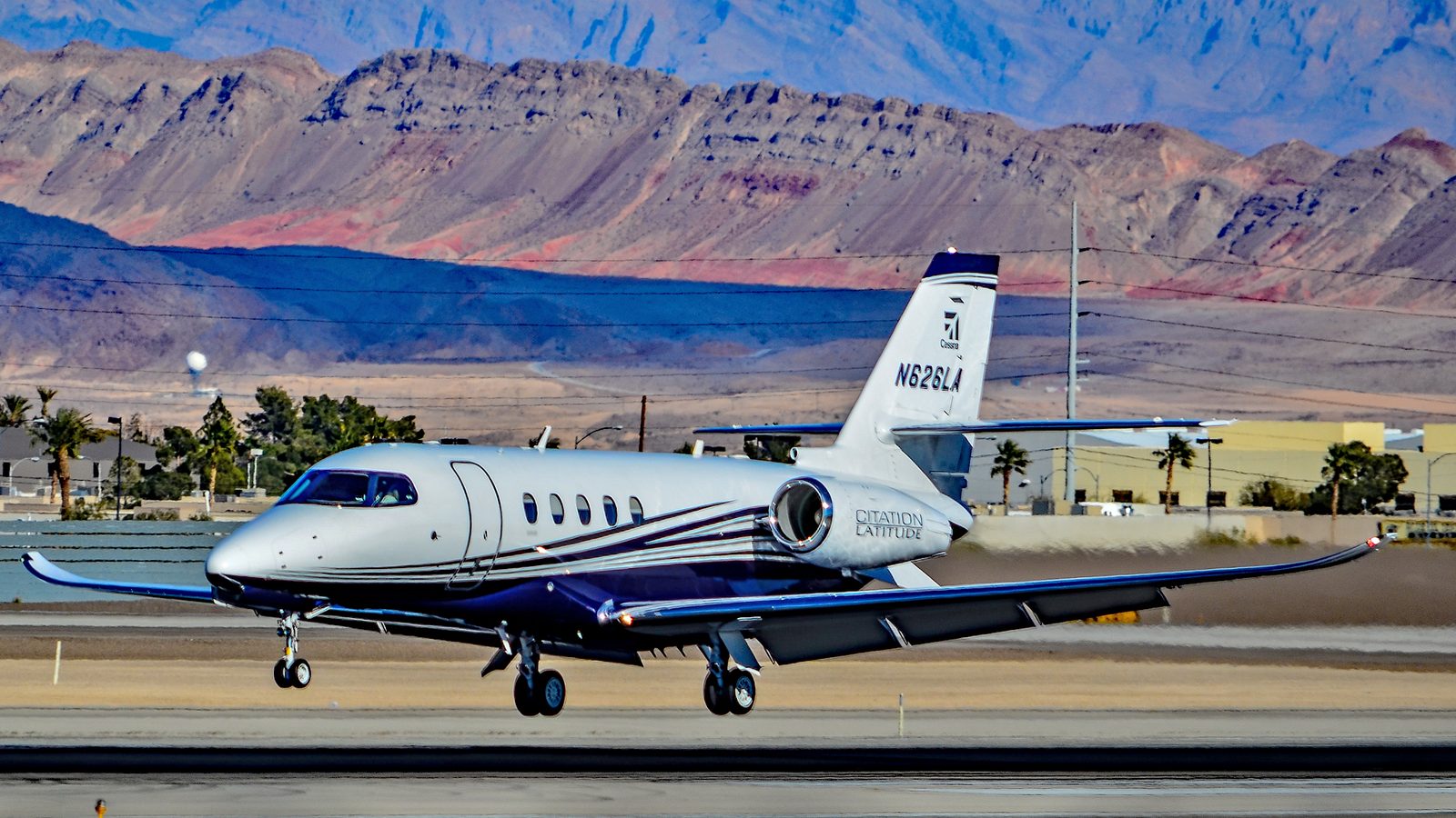 A silver and dark blue Cessna Citation Latitude lifting off, desert mountains behind, Citation Latitude titles on the engine